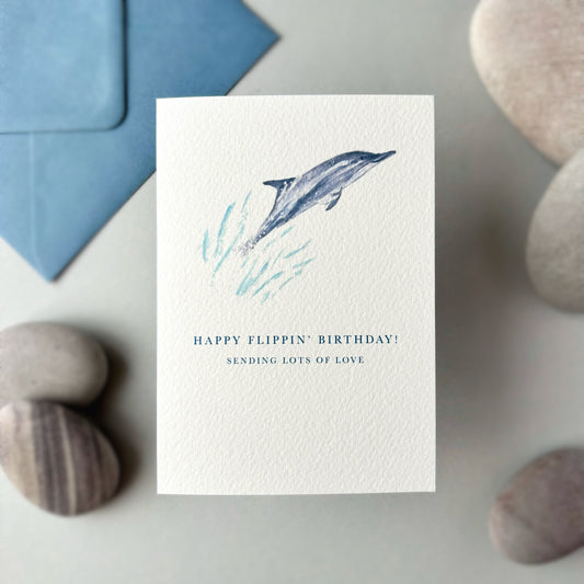 Greeting card with dolphin illustration and humorous birthday message, placed on a surface with stones and a blue envelope.