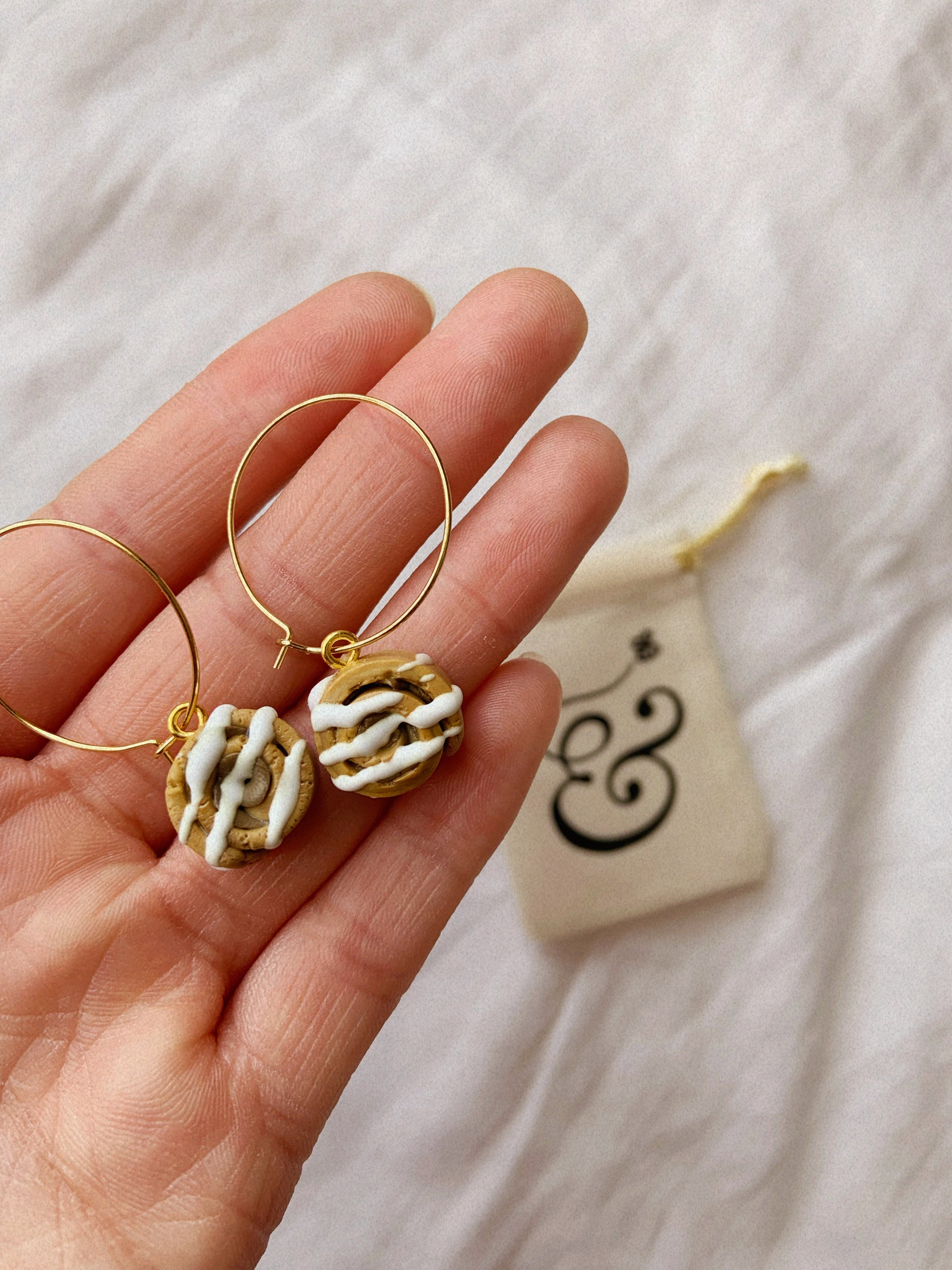 A hand holding a pair of cinnamon bun earrings, whilst a gift bag can be seen in the background.