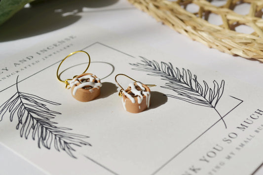 Miniature cinnamon buns made out of polymer clay. They have earrings components attached to them and are sitting on a Honey and Incense branded thank you card.