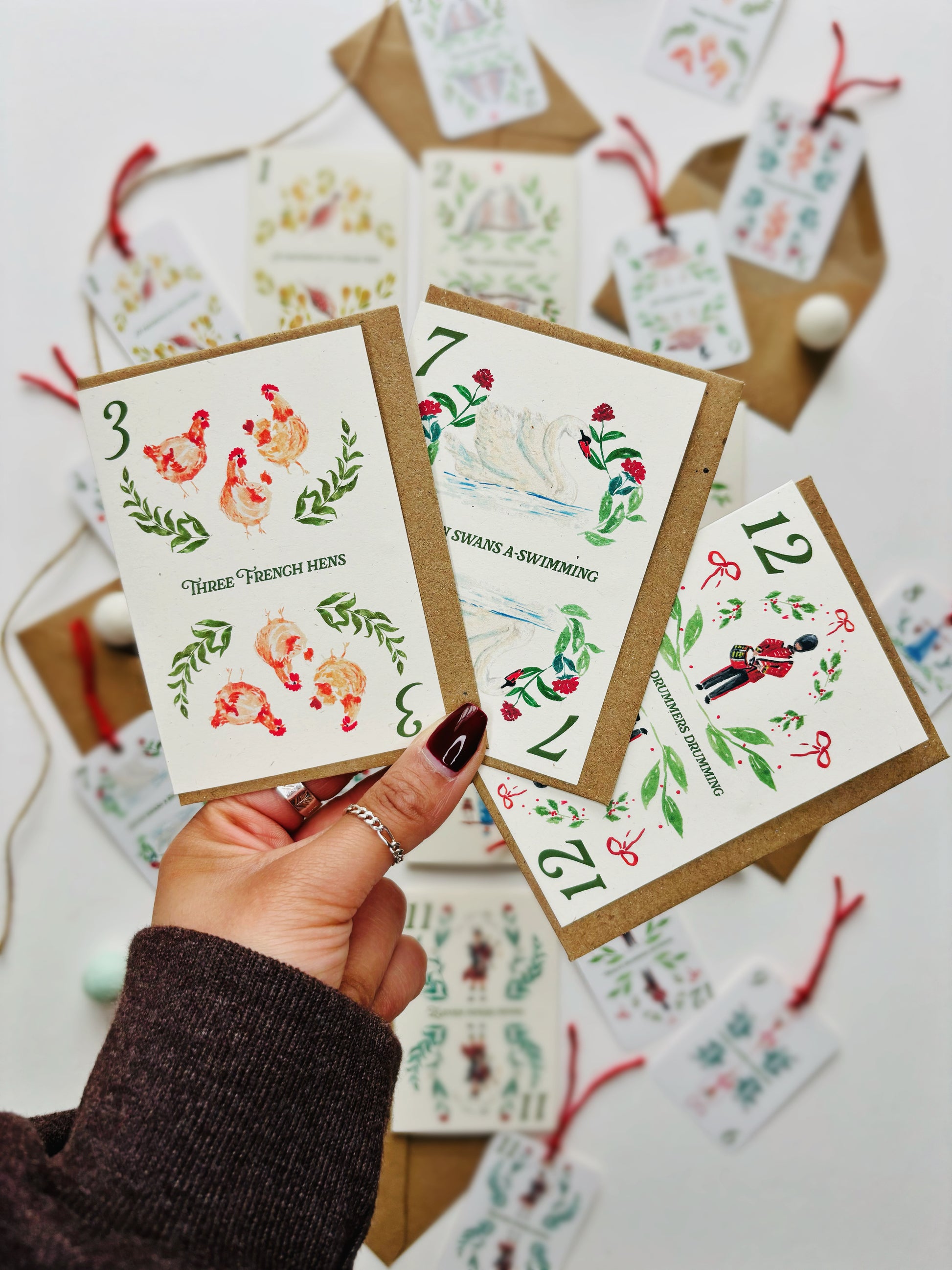 A hand holding 3 cards from a range of 12 days of Christmas cards, each featuring hand painted depictions of each of the 12 gifts from the famous carol