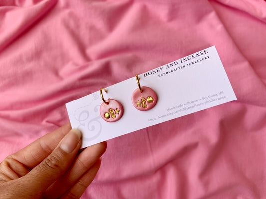 shimmery pink circles with gold seashells, starfish and seahorses on, on gold huggie hoop earrings.,