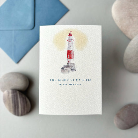 Greeting card with lighthouse illustration and birthday message on a stone surface with a blue envelope.