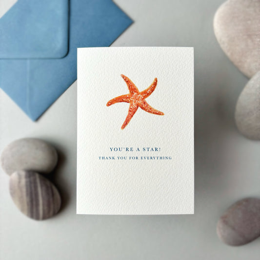 Card with a starfish design and text, placed on a surface with stones and a blue envelope.