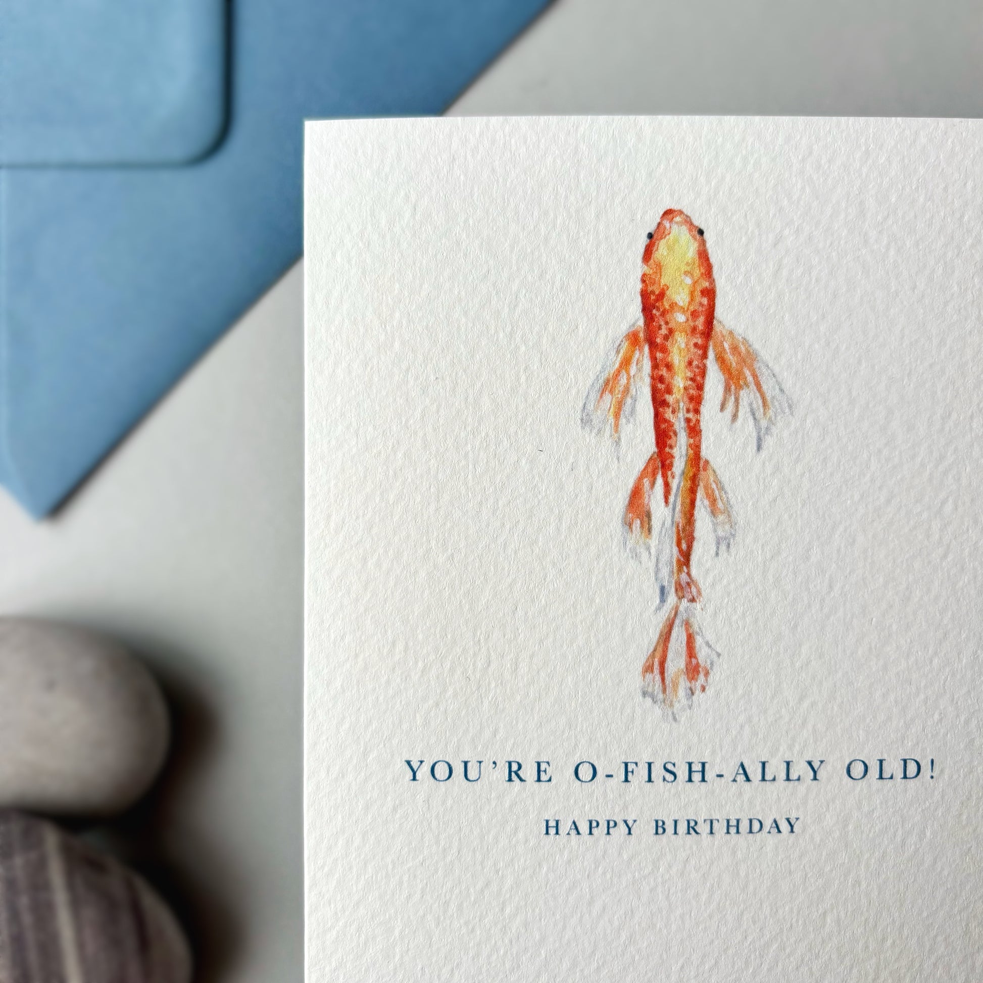 Birthday card with a koi fish illustration and humorous birthday message, placed on a surface with a blue envelope and stones.