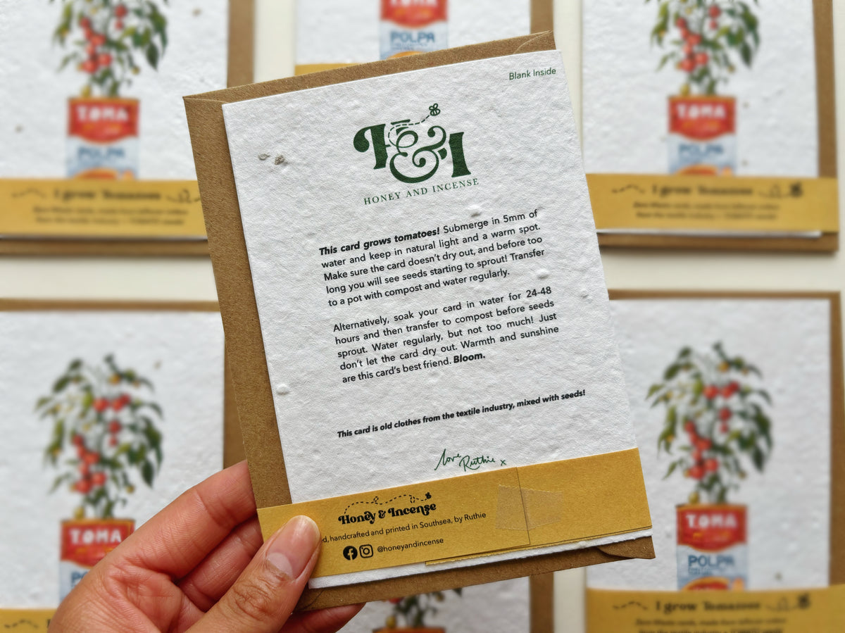 A hand holds a handmade tomato seeded card featuring a watercolour illustration of a tomato plant in a vintage red and blue tomato can, with text reading 'I grow tomatoes' and 'Zero waste seeds, made from flower petals from the tomato industry + TOMATO™ seed!'. Multiple matching cards are displayed in the background on a light wall.