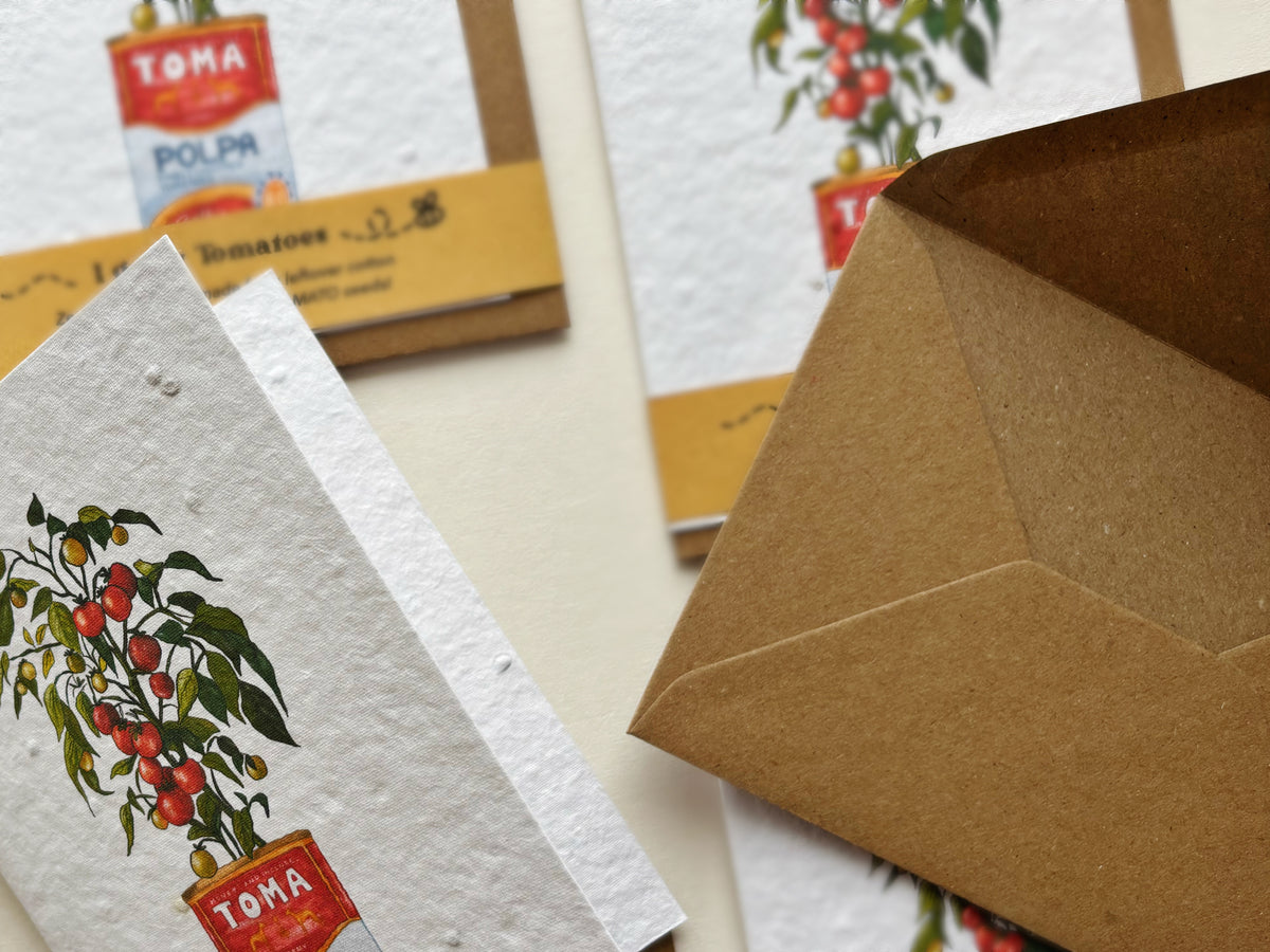 A close-up of a textured white seed paper card featuring a tomato plant illustration in a chopped tomato tin, alongside a brown kraft envelope on a light beige background.