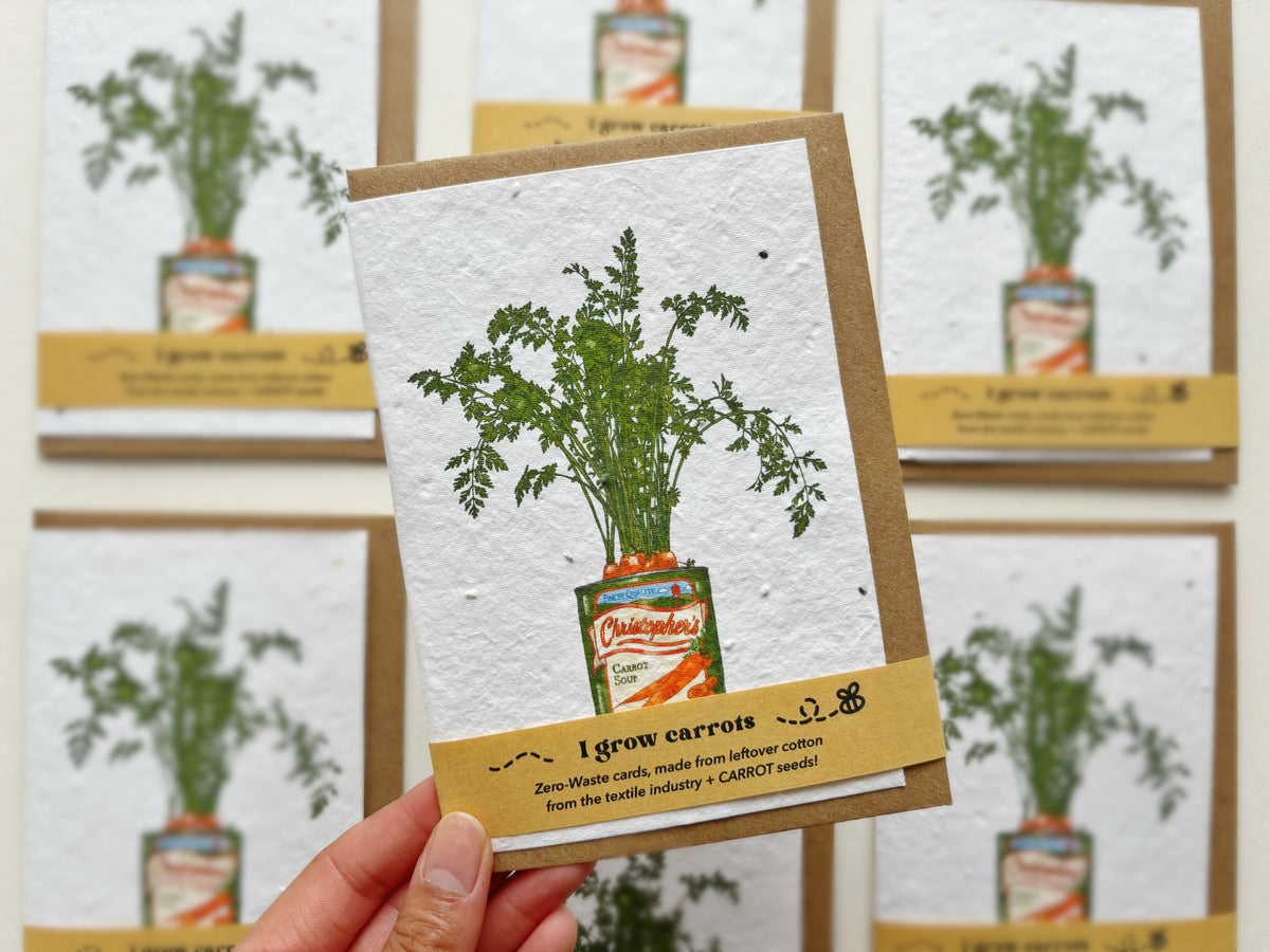 A handmade Plantable carrot seeded greeting card held by a hand, featuring an illustration of fresh green carrots growing out of a vintage can of carrot soup. The card has a brown kraft envelope behind it and a matching brown paper band at the bottom with the text 'I grow carrots' and a small graphic.