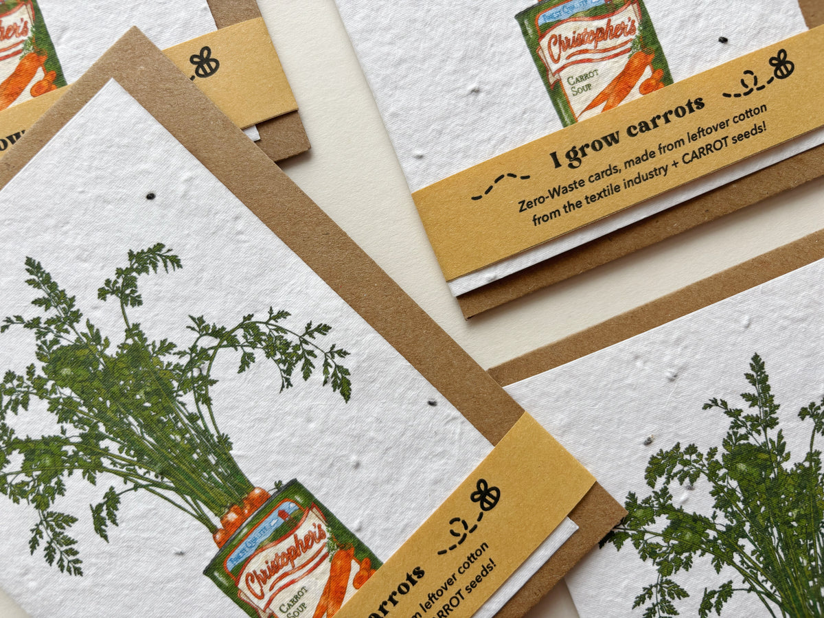 A close-up of several handmade carrot seed paper cards arranged on a light surface. The cards feature illustrations of carrots growing out of vintage carrot soup tins, kraft paper bands with text, and textured white paper backgrounds.