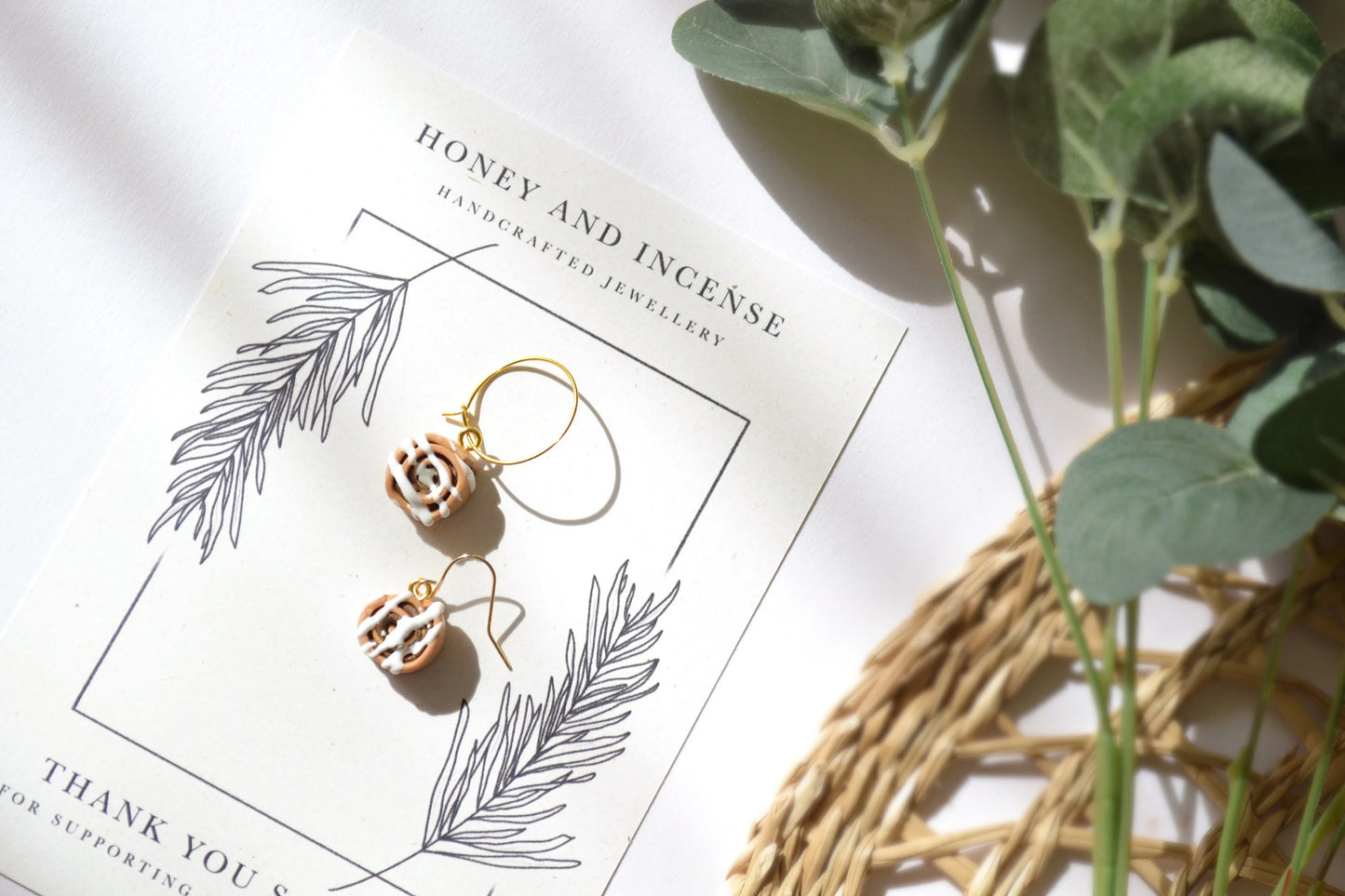 Small cinnamon bun earrings sitting on a Honey and Incense branded thank you note, with some eucalyptus leaves on the right hand side, framing the photograph.